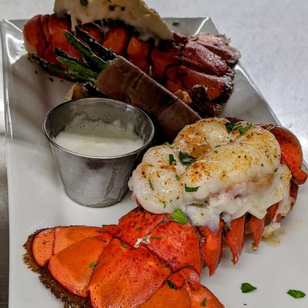 Two lobster tails, one stuffed with asparagus, served with a side of white sauce on a white plate.